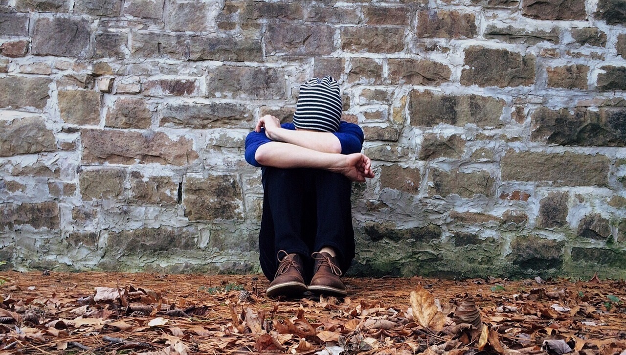 zdjęcie dziecka w pozycji siedzącej, zakrywającego twarz i chowającego głowę między kolanami. W tle ściana z cegieł